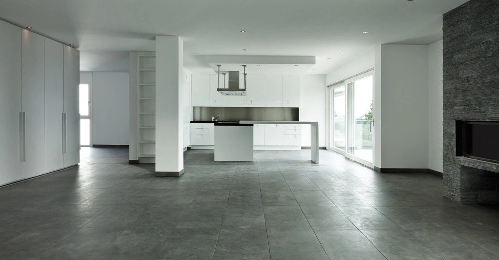 Helle moderne Küche mit weißen Schränken und Kücheninsel In an der Wand eingebaute Abzugshaube Großflächige Fenster und Schiebetüren lassen viel Licht in den Raum Graue Fliesen auf dem Boden.