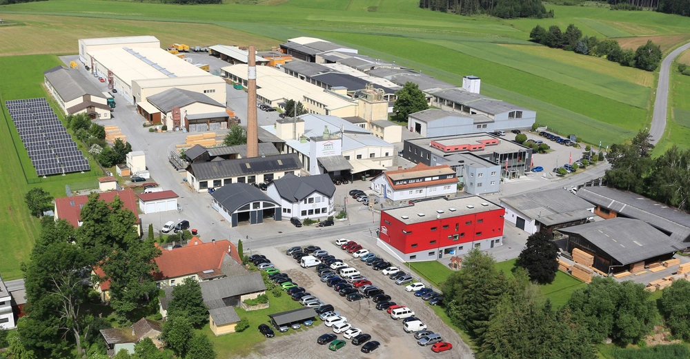 Mehrere Industriegebäude, umgeben von Feldern und Bäumen, mit einem Parkplatz voller Autos im Vordergrund. Eine Schornstein und Solarpanels sind sichtbar. Ländliche Umgebung mit Straßen und Wald.