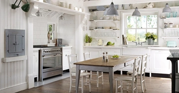 Ein rustikaler Küchentisch mit vier Stühlen steht im Zentrum einer hellen Küche mit weißen Schränken, Regalen voller Geschirr, und einem großen Fenster mit Blick ins Grüne. Links ein Herd.