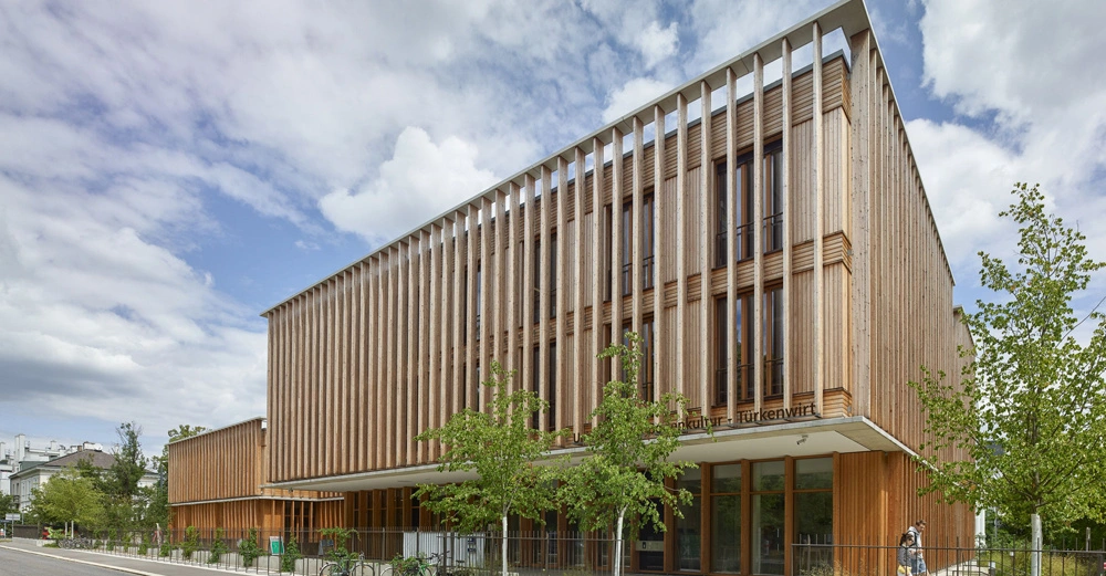 Modernes dreistöckiges Gebäude mit Holzfassade und vertikalen Lamellen Fenster. Stehende Bäume und Fahrräder im Vordergrund. Im städtischen Umfeld. Text: Verein für Baukultur - Türkenschanz.