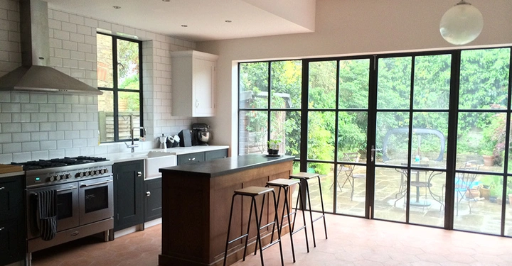 Galerie-Bild 5: Küche mit großem Herd und Dunstabzugshaube links, zentrale Kücheninsel mit Barhockern, große Fensterfront mit Blick auf einen begrünten Garten im Hintergrund.