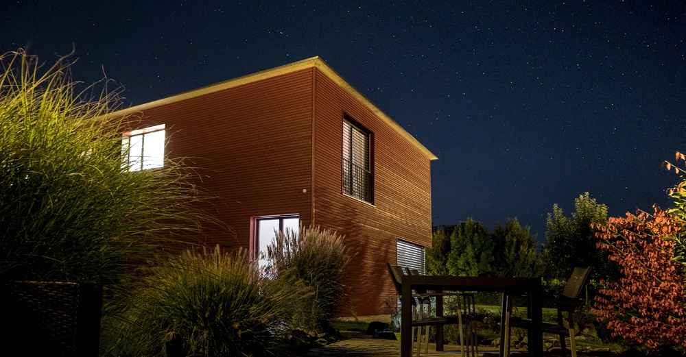 Rotes Haus leuchtet in der Nacht von innen im Garten mit Pflanzen und Sternenhimmel im Hintergrund