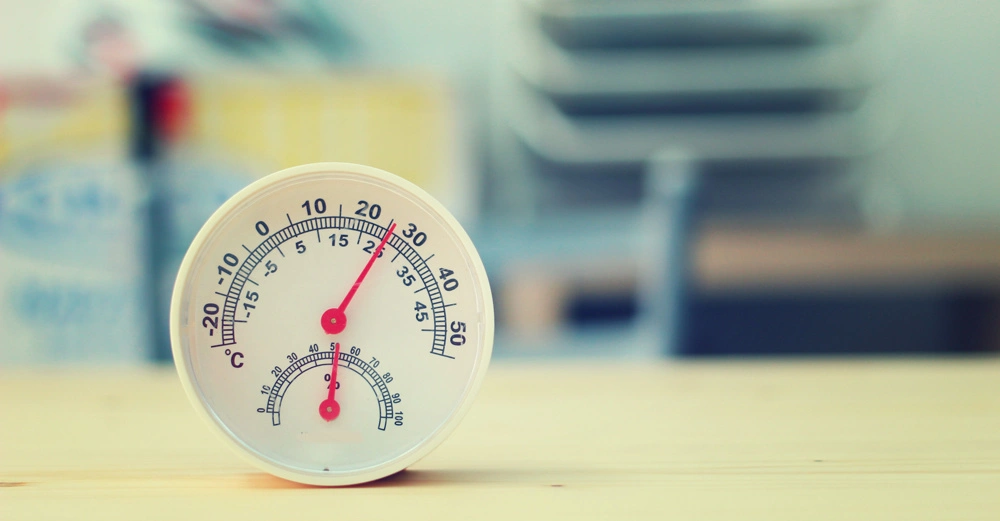 Ein kreisförmiges Thermometer zeigt 22 Grad Celsius an steht auf einem Holztisch in einem unscharfen Innenraum.