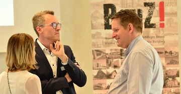 Drei Personen sprechen miteinander in einem Raum. Der Mann in der Mitte trägt einen Anzug und scheint nachdenklich. Im Hintergrund steht ein Plakat mit der Aufschrift Bauen und BauZ!