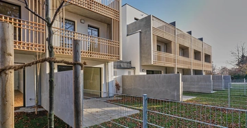 Mehrfamilienhaus mit Holzbalkonen und großen Fenstern umgeben von neu gepflanzten Bäumen und einem Metallzaun bei Tageslicht. Die Umgebung weist grüne Rasenflächen und herabgefallene Blätter auf.