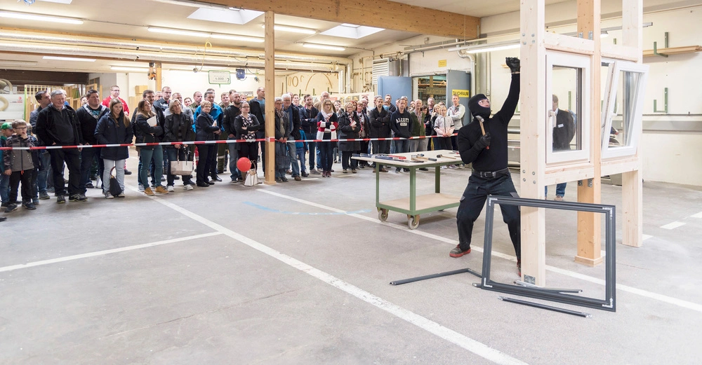 Ein Maskierter bricht mit Werkzeug ein Fenster ein in einer geräumigen Halle während eine Menge von Menschen hinter einer Absperrung zuschaut.