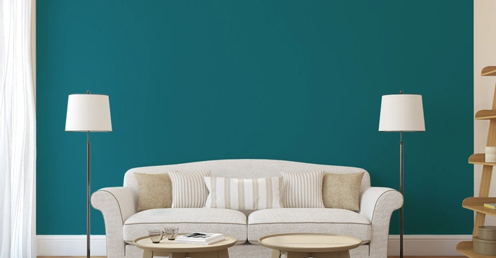 Beige Sofa stehend mit Kissen in einem Wohnzimmer beleuchtet von zwei Stehlampen mit einem türkisfarbenen Hintergrund und Holzmöbeln umgeben von Vorhängen und einem Teppich.