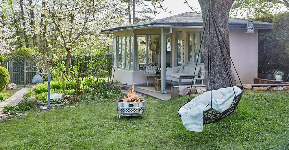 Hängesessel schwingt an einem Baum im blühenden Garten vor einem gemütlichen Haus Zimmer mit Glasfenstern Feuerstelle brennt davor Teppich liegt im Sessel Laternen hängen umher heller Tag.