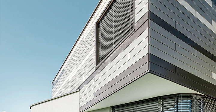 Modernes Gebäude mit gestreifter Fassade. Teilweise heruntergelassene Jalousien. Im Kontext eines klaren blauen Himmels und grüner Vegetation im Hintergrund.