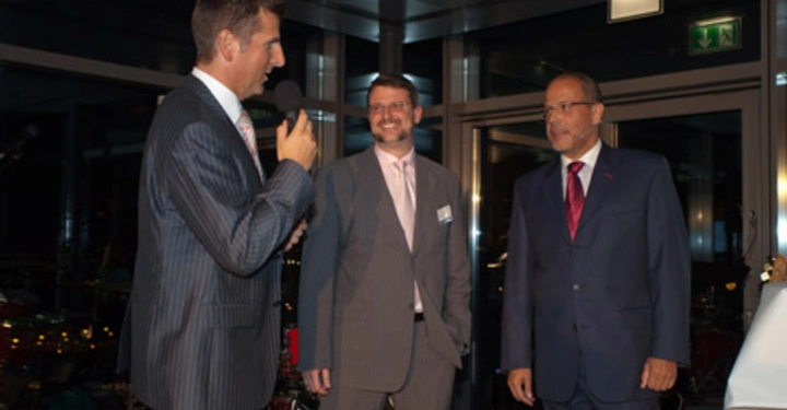 Galerie-Bild 70: Ein Mann spricht mit Mikrofon im Anzug links steht zwei weitere Männer im Anzug in der Mitte und rechts lachen beide über das Gesagte im Hintergrund ist ein modernes Gebäude bei Nacht