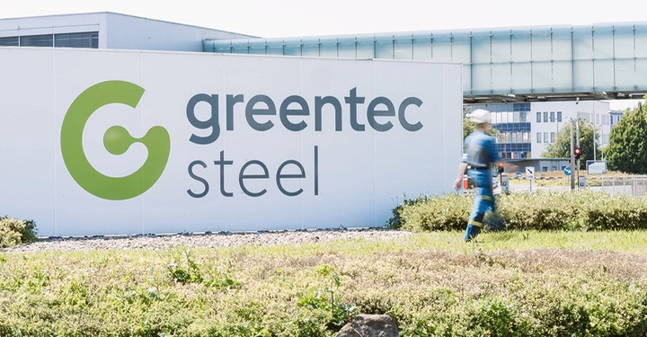 greentec steel Logo auf großer weißer Betonwand, vorne Rasen und Beete, ein Arbeiter geht vorbei