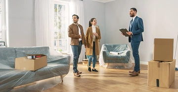 Drei Personen stehen in einem leeren, frisch renovierten Zimmer; zwei diskutieren mit einem Makler; Stühle sind mit Plastik bedeckt, Umzugskartons stehen auf dem Holzboden, großes Fenster im Hintergrund.