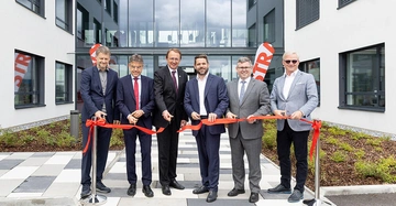 Sechs Männer schneiden ein rotes Band vor einem modernen Gebäude in einem festlichen Eröffnungszeremoniell. Im Hintergrund sind zwei STR-Banner und eine verglaste Brücke zwischen den Gebäuden zu sehen.