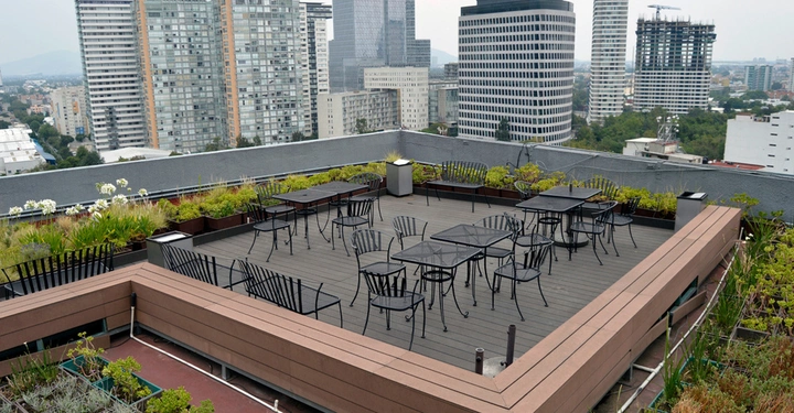 Dachterrasse mit dunklem Holzriemenboden und hellbrauner Attika, darauf stehen ein paar Gastrotische und Sessel. Im Hintergrund Skyline einer unbekannten Großstadt