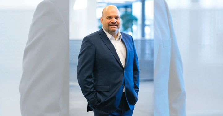 Galerie-Bild 4: Mann lächelt und trägt einen blauen Anzug mit Händen in den Taschen steht in modernem, lichtdurchflutetem Büro mit Glaswänden und Pflanzen im Hintergrund