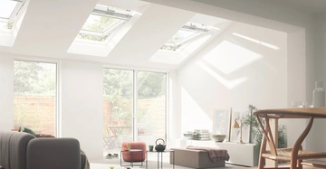 Graues Sofa steht in einem hellen Raum unter Dachfenstern im Sonnenlicht Wohnzimmer hat große Fenster Blick auf einen grünen Garten mit Pflanzen und Holzzaun ein Tisch mit Stuhl daneben.