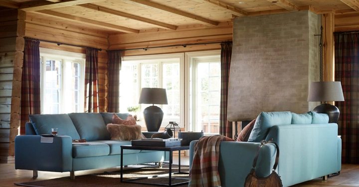 Hellblaue Sofas stehen in einem holzgetäfelten Wohnzimmer vor großen Fenstern ein Couchtisch dazwischen mit Deko und Lampen auf beiden Seiten ein gemauerter Kamin im Hintergrund.