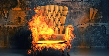 Ein großer, gepolsterter Sessel brennt heftig in einem heruntergekommenen Raum mit abblätternder Tapete und düsterer, beschädigter Wand.