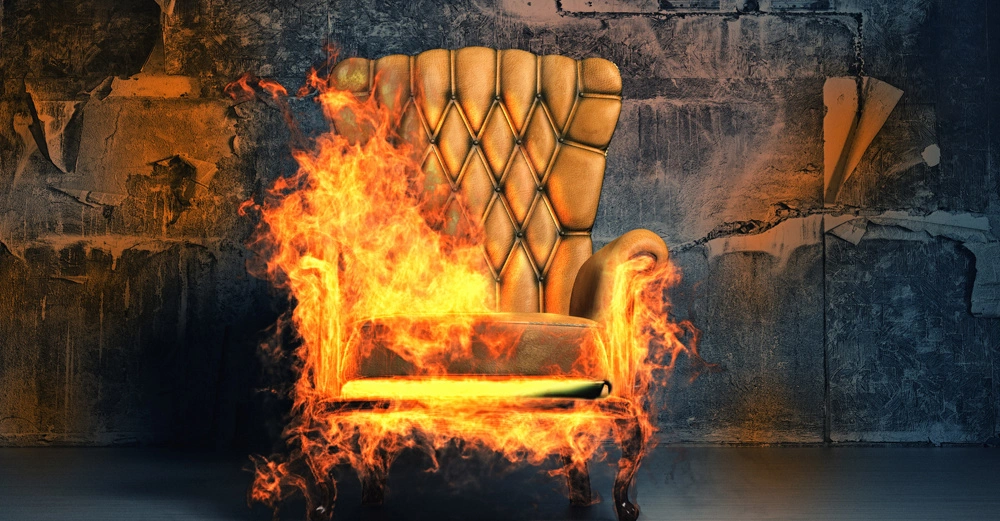 Ein großer, gepolsterter Sessel brennt heftig in einem heruntergekommenen Raum mit abblätternder Tapete und düsterer, beschädigter Wand.