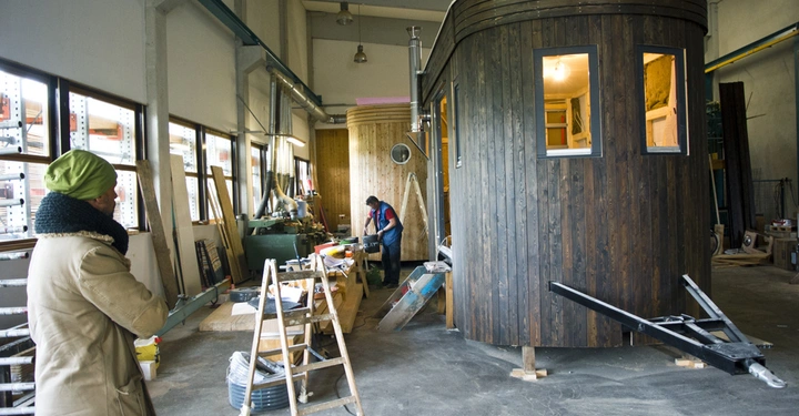 Galerie-Bild 10: Ein Holzhaus wird in einer Werkstatt gebaut, während ein Arbeiter es bearbeitet und eine Person zusieht. Die Umgebung ist voll mit Werkzeugen und Baumaterialien.