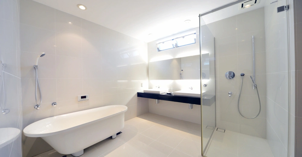 Eine freistehende Badewanne steht in einem weißen, modernen Badezimmer mit einer großen, verglasten Dusche, einem Doppelwaschbecken und Wandleuchten in einer hellen, minimalistischen Umgebung.