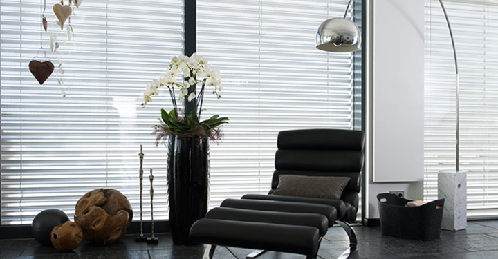 Galerie-Bild 3: Schwarzer Liegestuhl steht in einem modernen Wohnzimmer vor großen Jalousiefenstern eine Orchidee in einer Vase neben dekorativen Elementen wie Holzkugeln und einem hohen Bogenleuchte.