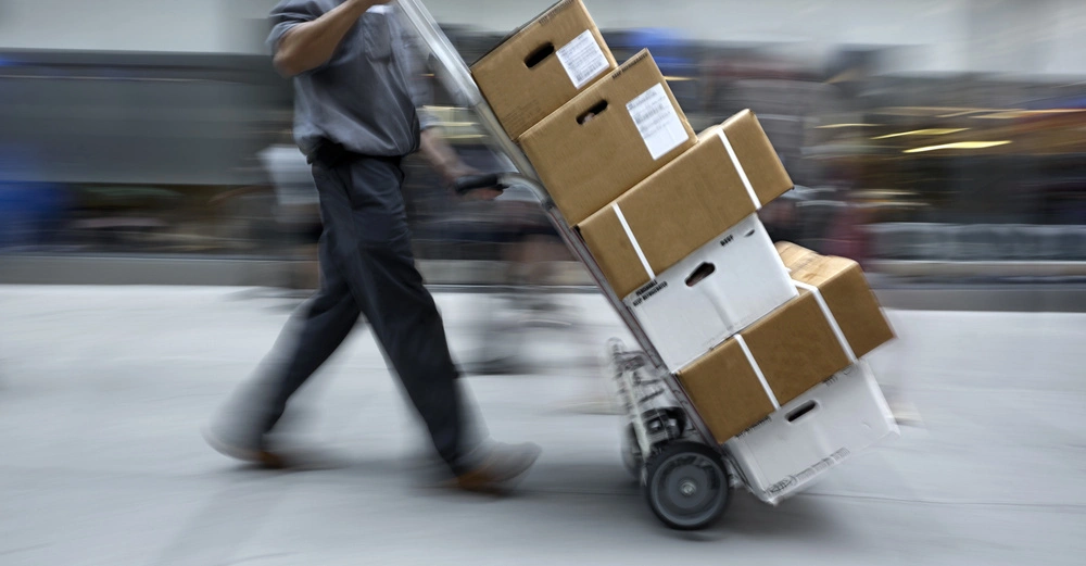 Ein Mann schiebt einen Handwagen mit gestapelten Kartons durch eine belebte Stadtstraße während des Tages