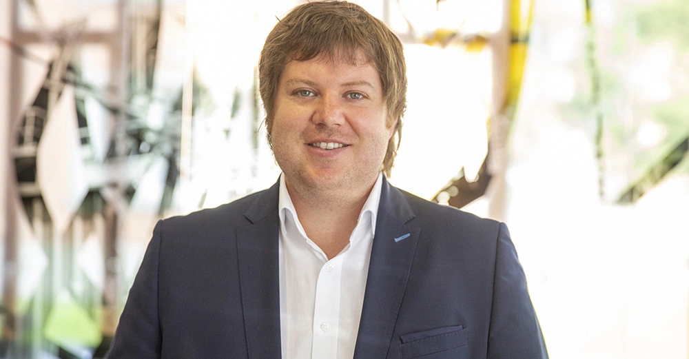 Mann lächelt trägt Anzug und Hemd steht vor unscharfem Hintergrund mit abstrakten Mustern und hellen Farben