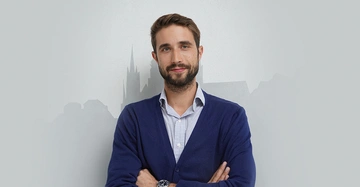 Portrait von R. Bergsleithner vor weißer Wand, verschränkte Arme, in weißem Hemd und blauer Weste, freundlich lächelnd