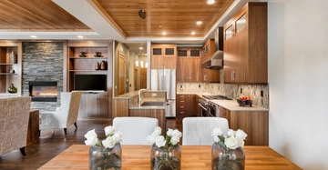 Holzverzierter Küchentisch mit Vasen und weißen Rosen steht im Vordergrund moderner Küche mit Holzmöbeln Edelstahlgeräten und Wohnzimmer im Hintergrund mit Kamin und eingebautem Flachbildfernseher.