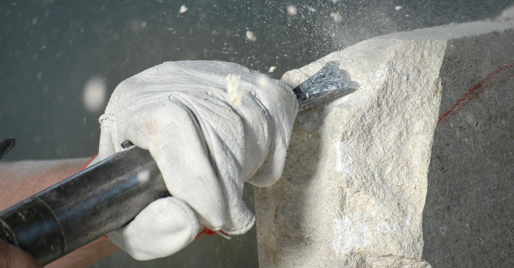 Ein Steinmetz bearbeitet ein Stück Naturstein mit einem Meißel. Kleine Bruchstücke fliegen dabei durch die Luft.