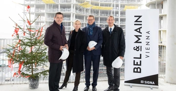 Vier Personen stehen mit Bauhelmen in der Hand vor einem Weihnachtsbaum und einem Bauprojekt mit einem Schild von "BEL & MAIN VIENNA SIGN.