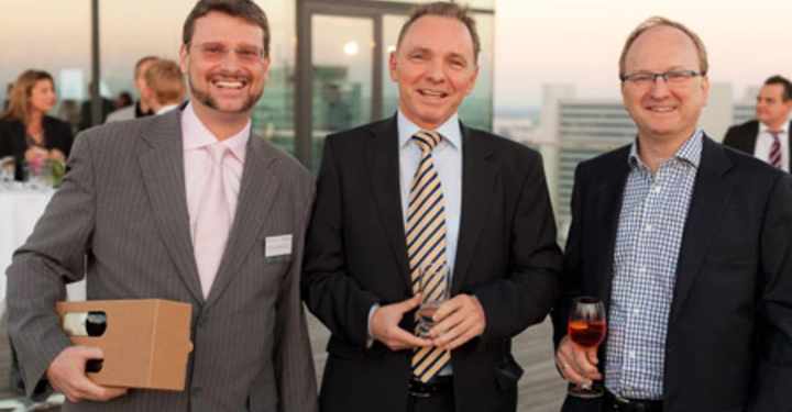 Galerie-Bild 38: Drei Männer stehen auf einer Terrasse und lächeln in die Kamera. Einer hält ein Geschenkpaket ein anderer ein Glas Wein. Im Hintergrund sind andere Gäste und ein städtisches Panorama zu sehen.