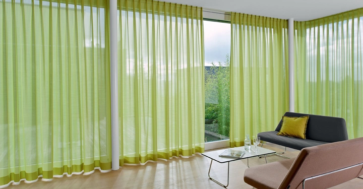 Galerie-Bild 8: Grünes Sofa und Sessel stehen in einem modernen Raum Sonnenlicht strömt durch bodenlange grüne Vorhänge und beleuchtet einen Holzboden mit einem Glastisch mit Wasserflasche und Magazin