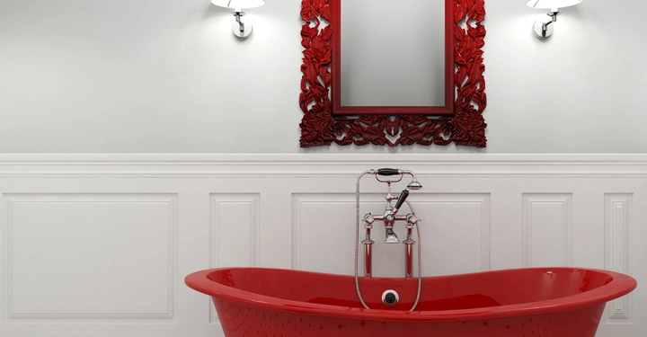 Rote freistehende Badewanne steht auf schwarz-weiß gefliestem Boden gefüllt mit Wasser. An der weißen Wand hängt ein roter kunstvoll verzierter Spiegel zwischen zwei Wandlampen.