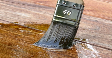 Ein Pinsel mit der Aufschrift 40 streicht braune Holzlasur auf eine hölzerne Oberfläche in einem gut beleuchteten Raum.