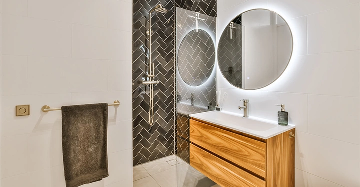 Spiegel hängt über Waschtisch im Badezimmer der von einer hölzernen Kommode unterstützt wird Dusche mit schwarzen Fliesen Handtuch hängt an goldener Stange weiße Wände und Bodenfliesen runden den Raum ab