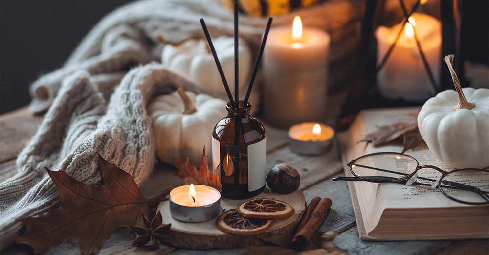 Braunes Glasfläschchen mit Duftstäbchen steht auf einem Holzbrett daneben brennende Teelichter getrocknete Orangenscheiben Kürbisse Zimtstangen Buch und Brille in gemütlicher Herbstszenerie drinnen.