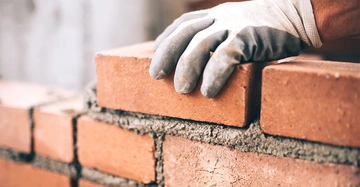 Hand trägt Arbeitshandschuh richtet eine Mauer aus roten Ziegelsteinen in einer Baustellenumgebung aus