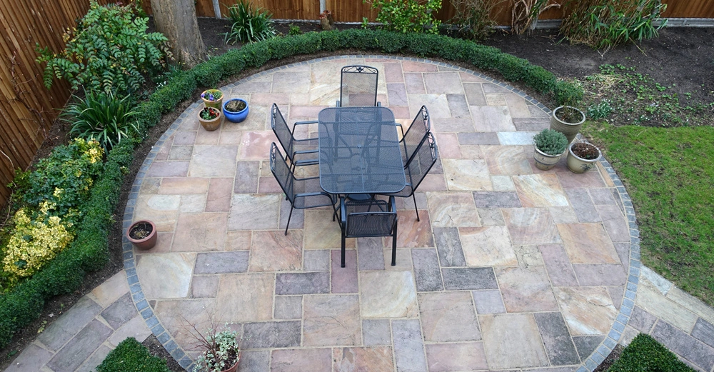 Metalltisch und Stühle auf einer gepflasterten Terrasse umgeben von Pflanzenkübeln und einer Hecke in einem Garten mit einer Holzlattenzaunumrandung.
