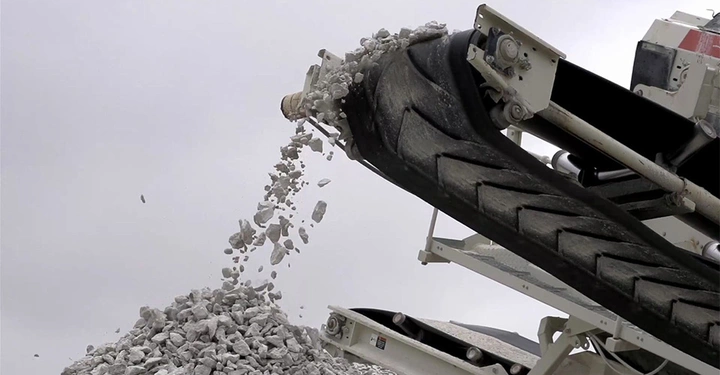 Ein laufendes Förderband befördert zerbrochene Steine und Sand nach unten in einen wachsenden Haufen. Hinter ihm ist ein grauverhangener Himmel.