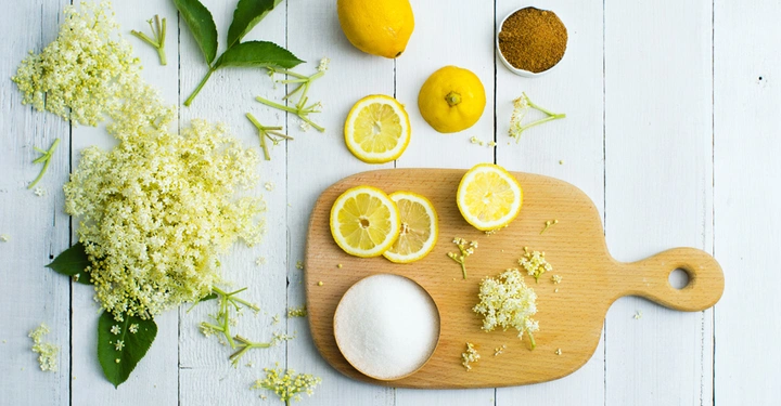 Holzbrett liegt auf einem weiß gestrichenen Holztisch Zucker Zitronenscheiben und Holunderblütenblätter liegen auf dem Brett daneben sind ganze Zitronen brauner Zucker und weitere Holunderblüten verteilt.