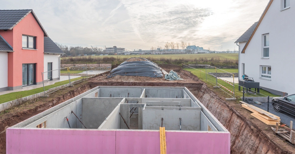 Offene Baugrube mit einem im Bau befindlichen Betonkeller, links uns rechts neben der Baugrube stehen neue Einfamilienhäuser