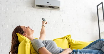 Frau liegt enspannt auf einem gelben Sofa in ihrer Wohnung und bedient mit einer Fernbedienung ihre Split Klimaanlage, die für angenehm kühle Raumluft sorgt.