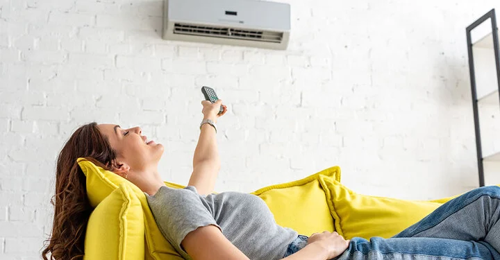 Frau liegt enspannt auf einem gelben Sofa in ihrer Wohnung und bedient mit einer Fernbedienung ihre Split Klimaanlage, die für angenehm kühle Raumluft sorgt.