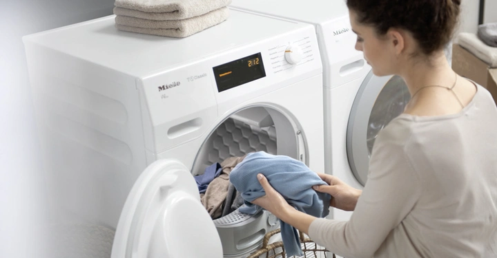 Eine Frau entnimmt Kleidung aus einem weißen Wäschetrockner der Marke Miele in einem hellen Hauswirtschaftsraum. Drei Handtücher liegen auf dem Trockner.