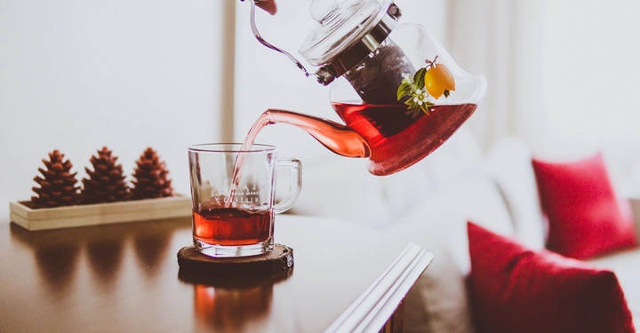Galerie-Bild 2: Eine Hand gießt roten Tee aus einer Glaskanne in eine Tasse auf einem Tisch vor einem Sofa mit roten Kissen und zwei Tannenzapfen im Hintergrund.