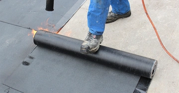 Eine Person in blauer Arbeitskleidung rollt ein brennendes schwarzes Bitumenband auf einen Betonboden aus um es zu versiegeln oumgeben von Werkzeugen und Materialien für Dacharbeiten