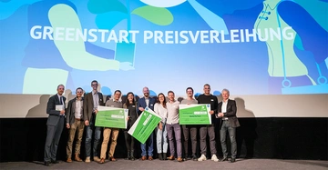 Gruppe von Menschen hält große symbolische Schecks, steht auf einer Bühne bei einer Preisverleihung. Im Hintergrund ein großer Bildschirm mit dem Text GREENSTART PREISVERLEIHUNG.