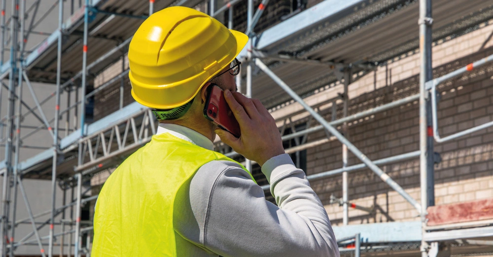 Mann mit gelbem Sicherheitshelm steht vor einem Baugerüst und hält sich sein Handy ans Ohr.